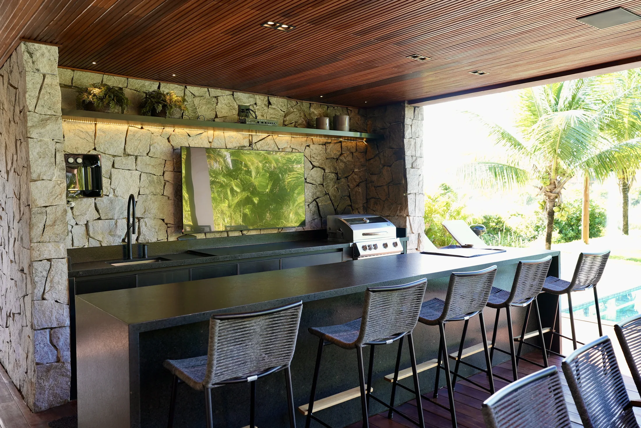 Bar e cozinha externa com bancada de pedra, banquetas e painel de pedra